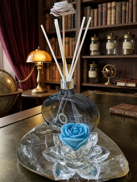Difusor de ambiente em vidro fumê com varetas brancas, flor de madeira e pingente prateado, ao lado de uma vela em formato de rosa azul sobre base de cristal em forma de lótus, em uma mesa de biblioteca