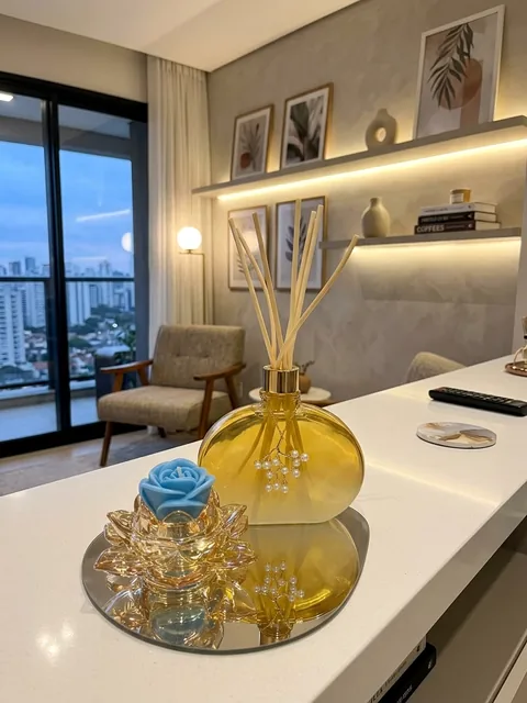 Difusor de ambiente em vidro âmbar com galho de pérolas e vela de rosa azul em lótus dourado, sobre bandeja espelhada em bancada de sala