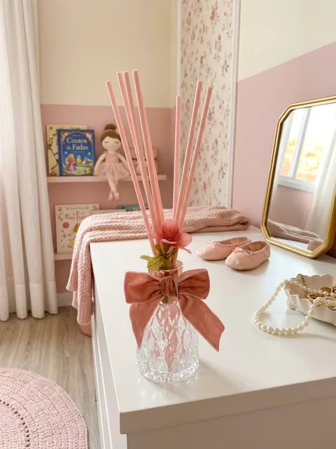 Difusor de ambiente em vidro lapidado com varetas rosa, flor e laço de veludo rosé, sobre cômoda branca em quarto infantil