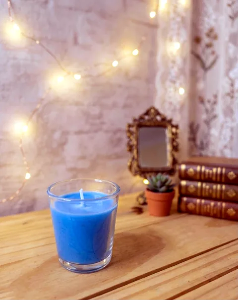 Vela azul em copo de vidro sobre mesa de madeira, com luzes de fada e espelho antigo ao fundo