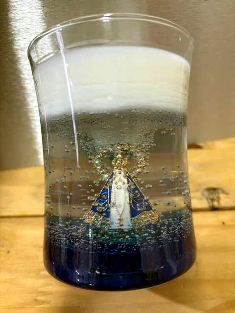 Vela decorativa em copo de vidro com camadas azul, transparente e branca, com medalha de Nossa Senhora Aparecida e pequenas bolhas