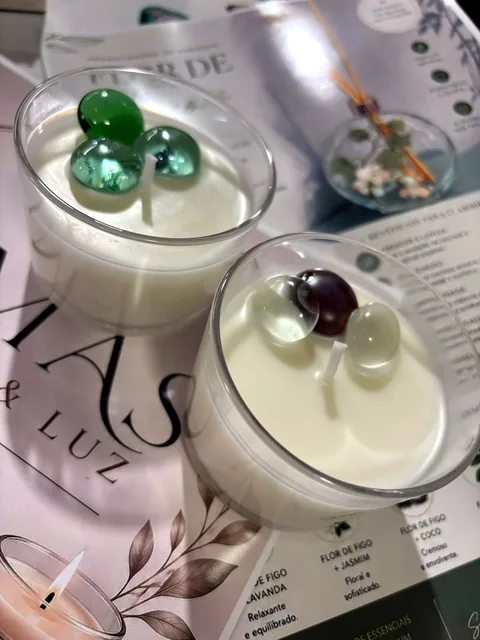 Duas velas de cera branca em copos de vidro, decoradas com pedras de vidro verdes, transparentes e marrom, sobre um folheto da marca